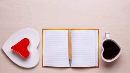 Coffee cup jelly cake in form of heart and paper blank notebook with pen on desk, top view copy space for textの写真素材