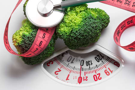 Diet healthy eating weight control concept. Closeup green broccoli with measuring tape and stethoscope on white scalesの写真素材