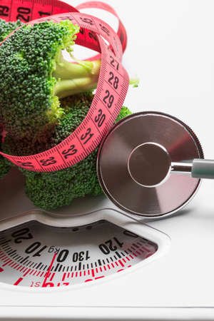 Diet healthy eating weight control concept. Closeup green broccoli with measuring tape and stethoscope on white scalesの写真素材