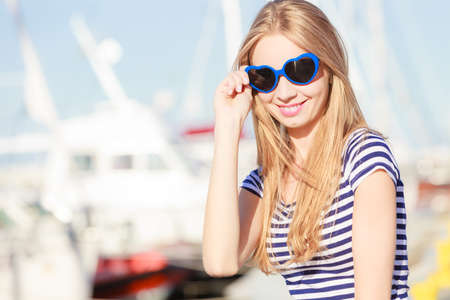 Travel tourism and people concept. Fashion blonde girl with blue heart shaped sunglasses in marina against yachts in portの写真素材