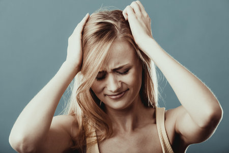 Negative human emotions, facial expressions, reaction attitude. Closeup stressed worried young woman having really bad headache hurt pain holding head with hands on blue grayの写真素材