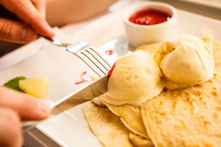 Human hands at dinner table holding fork and knife eating pancake with ice cream and strawberry sauceの写真素材