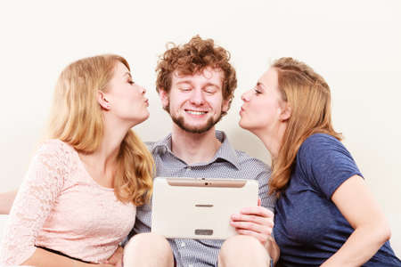 Young women girls kissing handsome man guy with tablet. Happy friends having fun relaxing at home.の写真素材