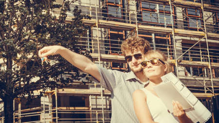 Real estate and family concept - young couple on front of new big modern house with blueprint project and key dreaming about homeの写真素材