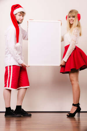Couple woman and man holding empty blank banner with copy space. Pretty girl in earmuffs and handsome man wearing santa claus hat studio on gray. Christmas xmas advertisement.の写真素材
