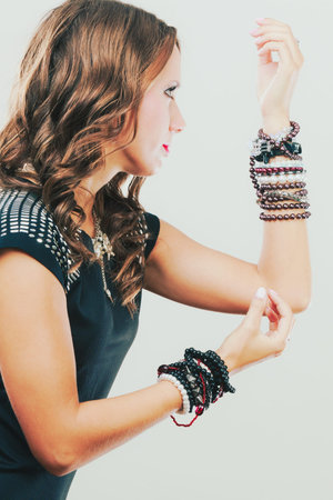 Pretty young woman wearing multiple bracelets jewellery necklace in black elegant evening dress side view on greyの写真素材