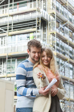 Real estate and family concept - young couple on front of new big modern house construction site with keys dreaming about homeの写真素材