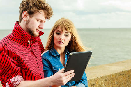 Modern technology devices and tourism concept. Young couple outdoor against sea water with tablet enjoying summer holidaysの写真素材