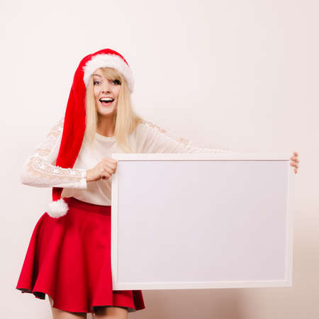 Happy woman in santa helper hat holding empty blank banner with copy space. Pretty girl in studio on gray. Christmas xmas advertisement.の写真素材