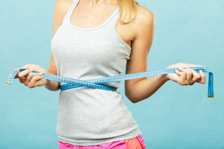 Weight loss, slim body, healthy lifestyle concept. Fit fitness girl measuring her waistline with measure tape on blueの写真素材