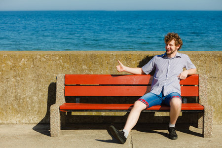 Happy smiling young man sitting on bench by sea ocean water outdoors. Summer vacation.の写真素材