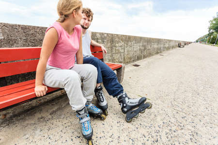 Young people friends in training suit with roller skates. Woman and man relaxing on bench outdoor.の写真素材