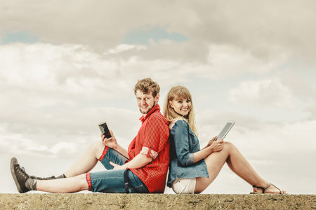 Modern technology devices. Young couple outdoor against sky man with smartphone, woman with tablet enjoying summer holidayの写真素材