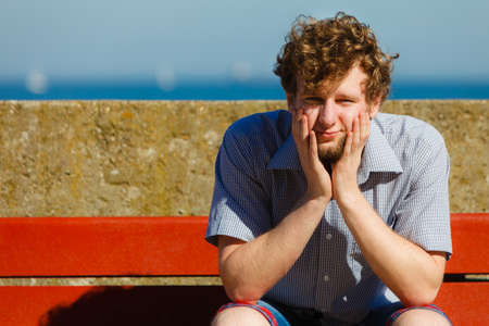 Tired exhausted man sitting on bench by sea ocean. Young guy relaxing outdoor. Summer vacation.の写真素材