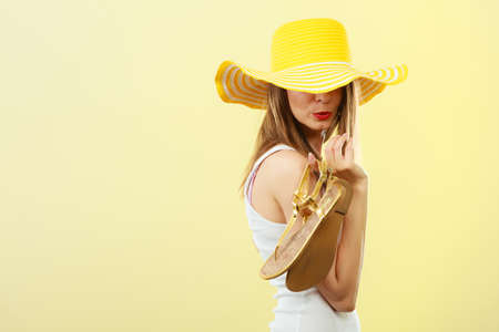 Holidays summer fashion concept. Woman in big yellow hat holding golden sandals in hand bright background.の写真素材
