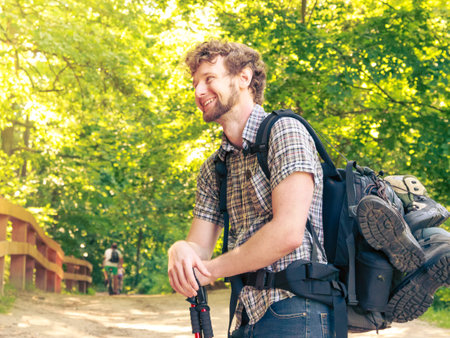Adventure, tourism, enjoying summer time - young tourist man with backpack sticks hiking in forest...