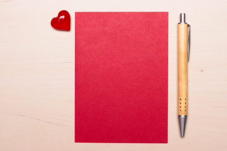 Red blank sheet of paper with heart love symbol and pen on wooden surface. Background for valentines...