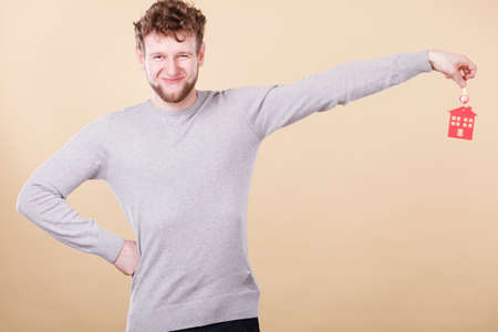 Young bearded smiling man holding new house key with red home shape. Real estate and housing concept.の写真素材