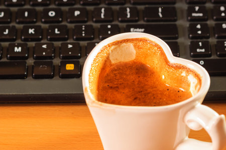 Heart shaped mug cup of coffee next to laptop notebook computer keyboard.の写真素材