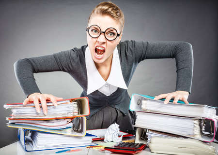 Job work frustration bureau paperwork workaholism concept. Upset secretary at her desk. Young office lady in glasses expressing anger.の写真素材