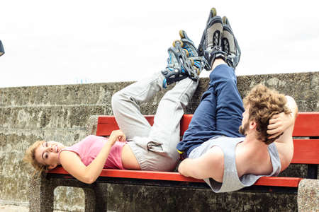 Young people friends in training suit with roller skates. Woman and man relaxing lying on bench outdoor.の写真素材