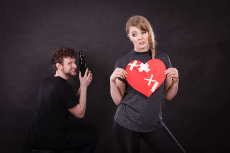 Unhappy couple. Family and alcoholism problems. Addiction and trouble of drinking. Man with alcohol bottle. Sad woman holds paper broken heart.の写真素材