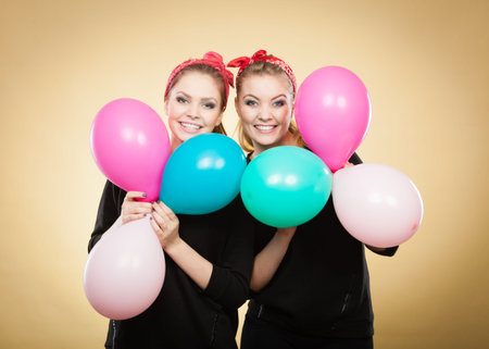 Preparation to birthday party. Two lovely happy retro girls preparing celebration. Smiling joyful women with multicolored balloons.の写真素材
