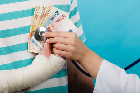 Corruption in medicine. Female doctor with stethoscope checking heart beating. Male bandaged hand with banknotes euro money.の写真素材