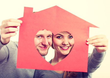 Love romance feelings ownership property future concept. Young cheerful couple holding house. Smiling girl and boy hugging showing red home with heart cutout.の写真素材