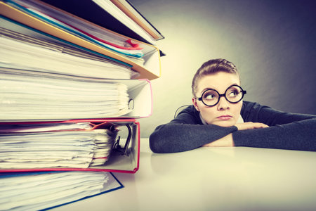 A lot of work and overwork concept. Terrified young businesswoman secretary with huge stack of binders. Scared accountant surrounded of to much work to do.の写真素材