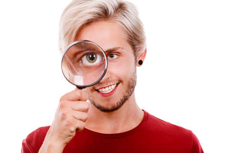 Investigation exploration education concept. Young smiling man holding on eye magnifying glass looking through loupe, isolated on whiteの写真素材