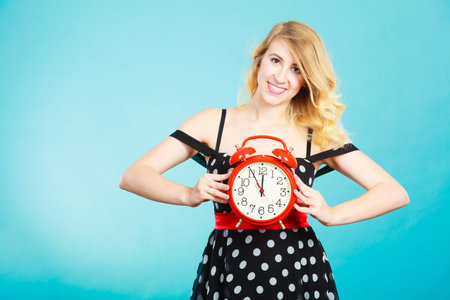 Management time concept. Blonde fashion girl wearing black dotted dress smiling face expression with alarm clock on blue.の写真素材