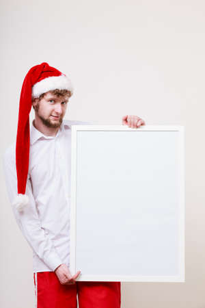 Man in santa claus hat holding empty blank banner with copy space. Handsome guy in studio on gray. Christmas xmas advertisement.の写真素材
