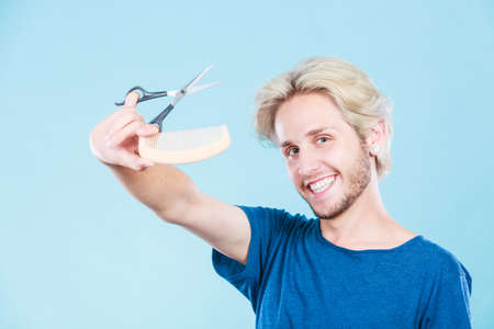 Style and fashion. Young trendy male hairstylist barber with new idea of look changing. Blonde guy with scissors and wooden comb creating hairdo coiffure, on blueの写真素材