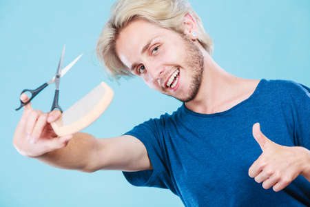 Style and fashion. Young trendy male hairstylist barber with new idea of look changing. Blonde guy with scissors and wooden comb creating hairdo coiffure, on blueの写真素材