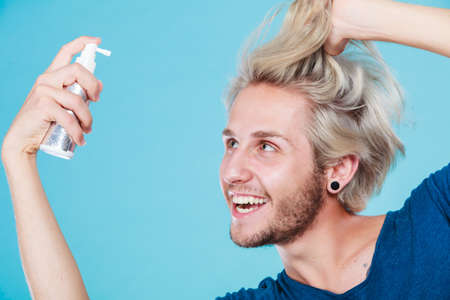 Hairdo haircare and makes hairstyle concept. Handsome young guy, fashion blonde metrosexual model applying spray cosmetic to his hairの写真素材