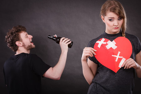 Unhappy couple. Family and alcoholism problems. Addiction and trouble of drinking. Man with alcohol bottle. Sad woman holds paper broken heart.の写真素材