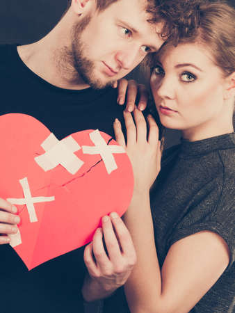 Broken heart difficult love concept. Sad unhappy couple woman and man holding paper red heart fixed with plaster bandage. Rift in relations.の写真素材