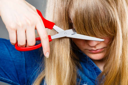 Little look change concept. Young blonde woman cutting down shearing her fringe short front hair. Girl using big scissors.の写真素材