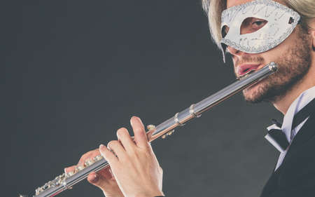 Holidays, people and celebration concept. Elegant young guy wearing suit white shirt bow tie and carnival venetian mask playing flute instrument on darkの写真素材
