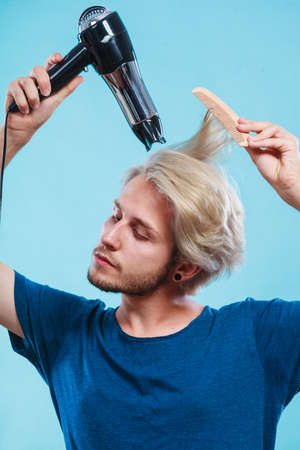 Style and fashion. Young trendy male hairstylist barber with new idea of look changing. Blonde man holding hair dryer and comb creating new hairdo, on blueの写真素材