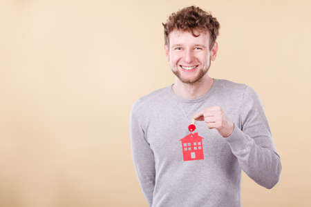 Young bearded smiling man holding new house key with red home shape. Real estate and housing concept.の写真素材