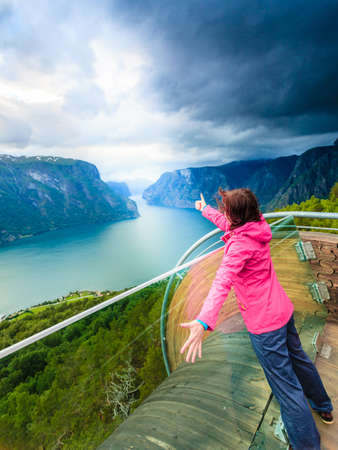 Tourist woman on Stegastein viewpoint enjoying Aurland fjord view making thumb up gesture sign, Sogn Og Fjordane, Norwayの写真素材