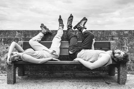 Young tired people friends in training suit with roller skates. Woman and man relaxing lying on bench outdoor.の写真素材