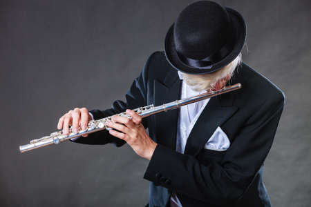 Classical music, passion and hobby concept. Elegantly dressed musician man playing on flute wearing black fedora hat. Studio shot on dark grey backgroundの写真素材