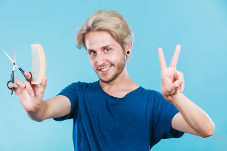 Style and fashion. Young trendy male hairstylist barber with new idea of look changing. Blonde guy with scissors and wooden comb creating hairdo coiffure, on blueの写真素材