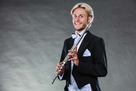 Classical music, passion and hobby concept. Portrait of elegantly dressed musician blonde young man holding flute. Studio shot on dark grey backgroundの写真素材
