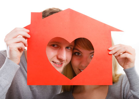 Love romance feelings ownership property future concept. Young cheerful couple holding house. Smiling girl and boy hugging showing red home with heart cutout.の写真素材