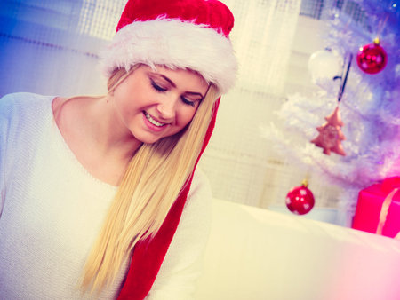 Seasonal concept. Happy blonde woman wearing santa hat, white christmas tree in background, indoor shot.の写真素材
