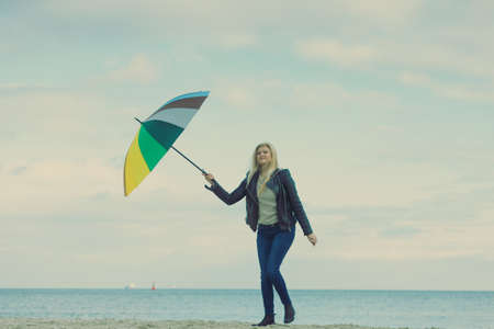 Happiness, enjoying cold autumn weather, feeling great concept. Woman holding colorful umbrella on beach near sea, sunny day and clear blue skyの写真素材